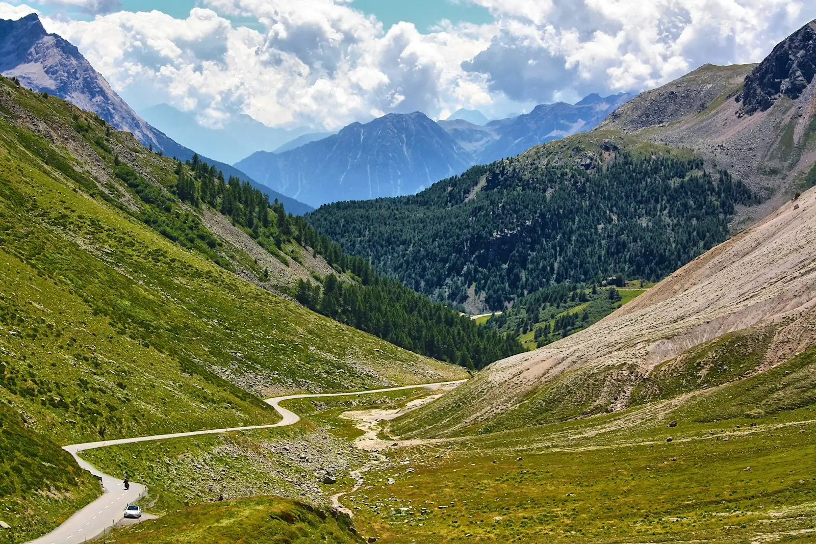 Route de montagne serpentant au fond d'une vallée alpine, sommets et nuages à l'horizon